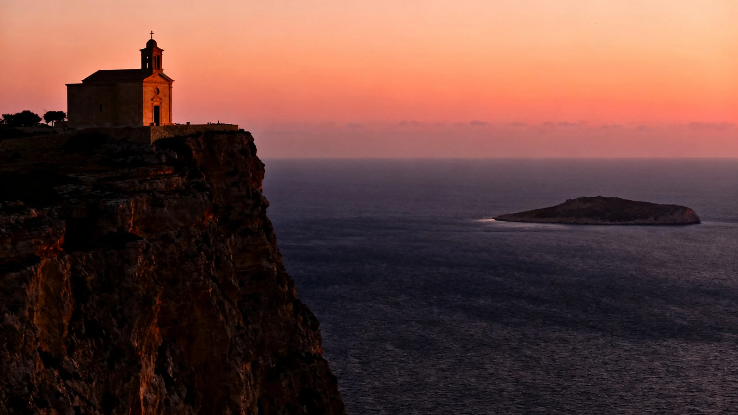 The Dingli Cliffs at sunset, with the chapel of St Mary Magdalene silhouetted against the orange sky and the cliff dropping to the dark Mediterranean below