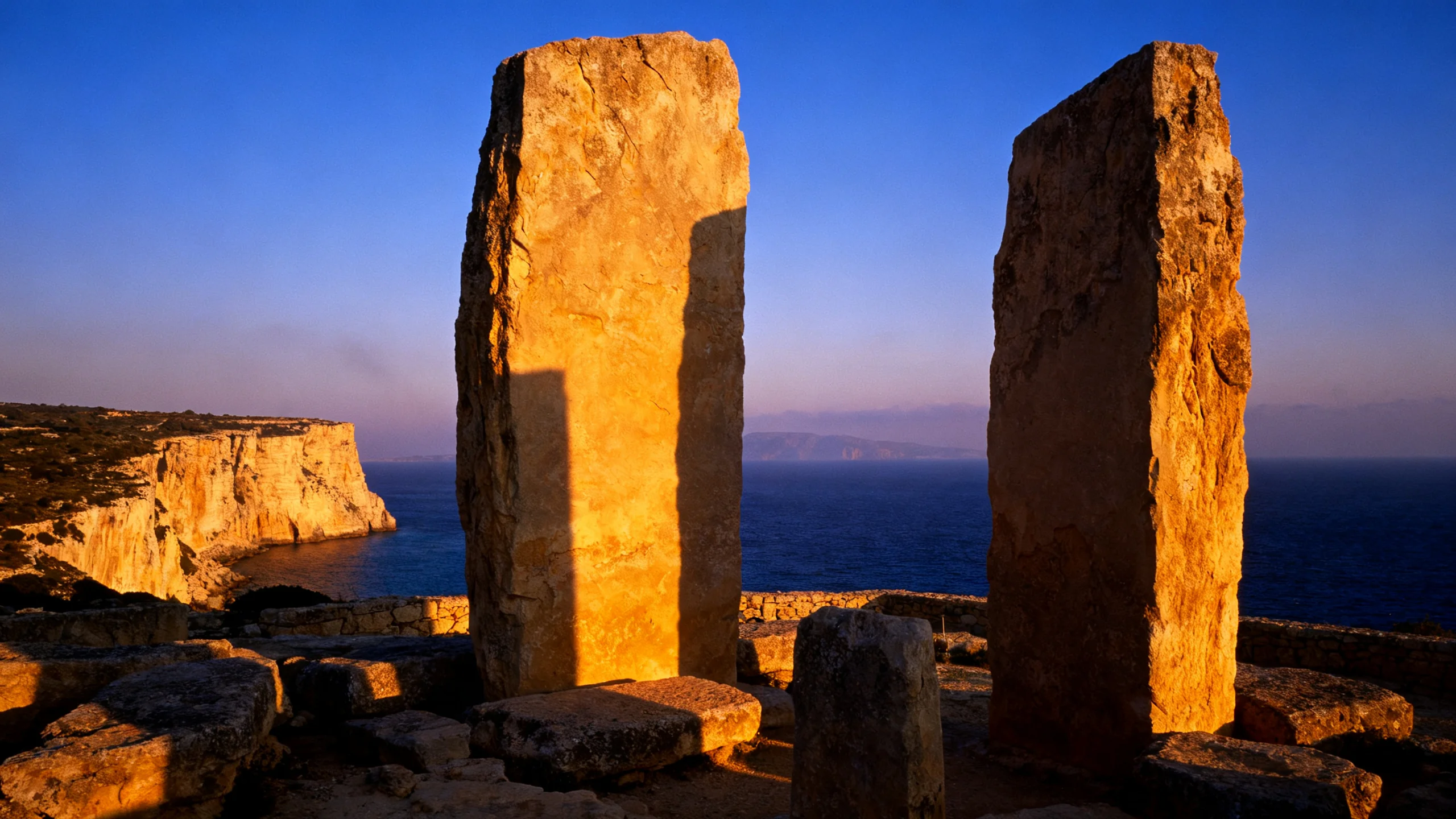 The Mnajdra temple complex at dusk, the upright limestone slabs catching the last warm light against the dark Mediterranean behind