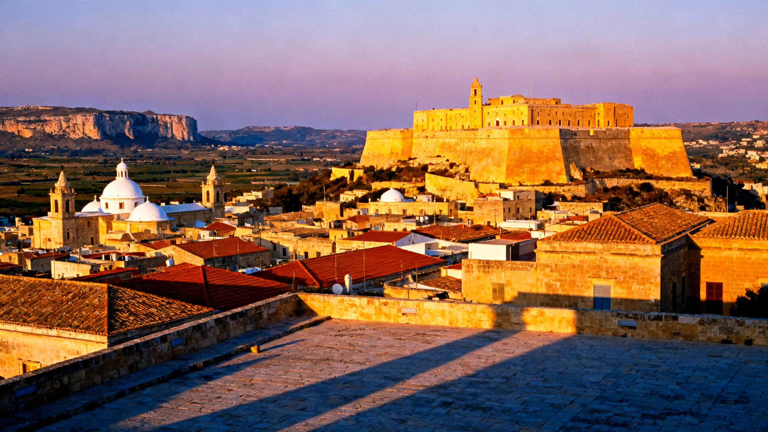 The Cittadella in Victoria, Gozo, rising above the rooftops at golden hour, with the central Gozo countryside stretching toward the western cliffs