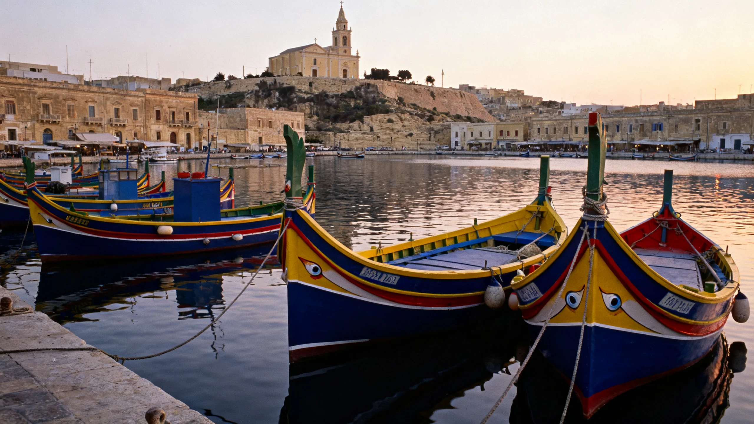 Painted luzzu boats in Marsaxlokk harbour at dawn, with the parish church of Our Lady of Pompei visible behind